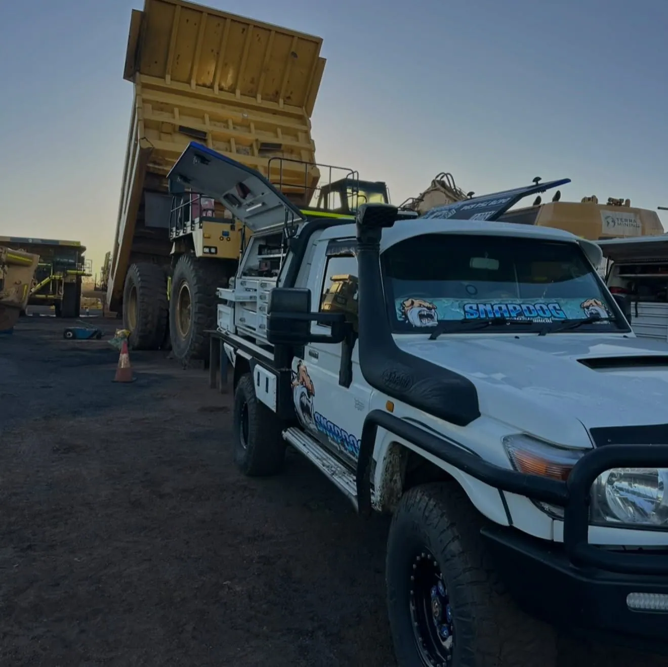 SnapDog's white Land Cruiser tray-back parked beside a large yellow dump truck at a mine site in late afternoon light.