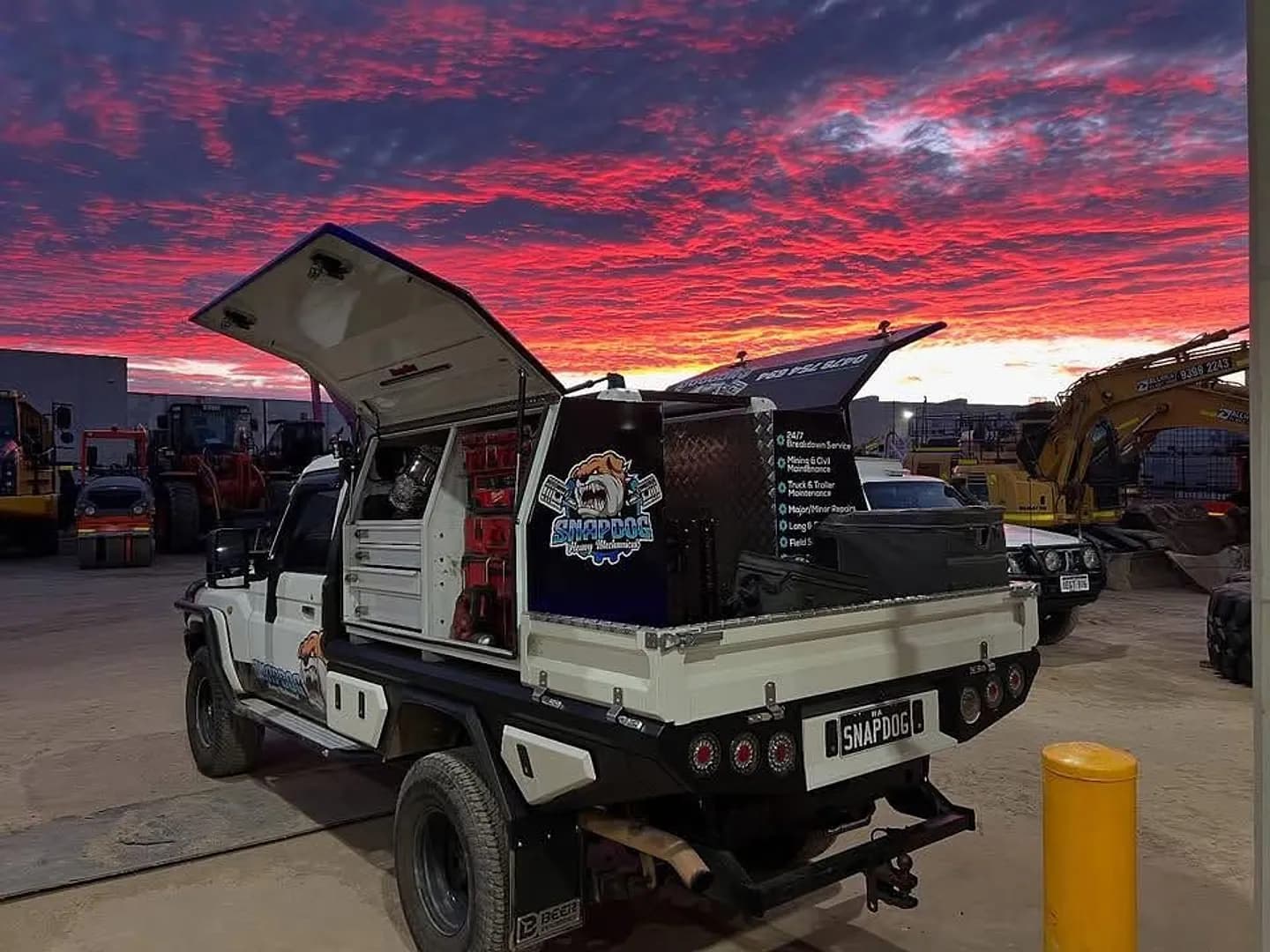 SnapDog's tray-back with its canopy doors raised against a red dusk sky, with excavators and loaders parked behind.
