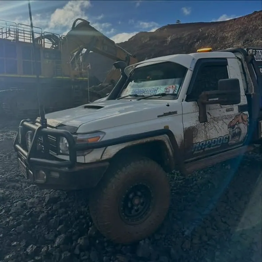 SnapDog's tray-back parked on rough ground below a mine pit wall with an excavator behind and low sun flaring across the lens.